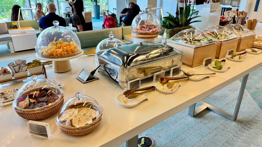 Mesa de buffet com queijos, frios, saladas, pães e chips em recipientes de vidro e madeira, com talheres de servir. Pessoas desfocadas ao fundo.
