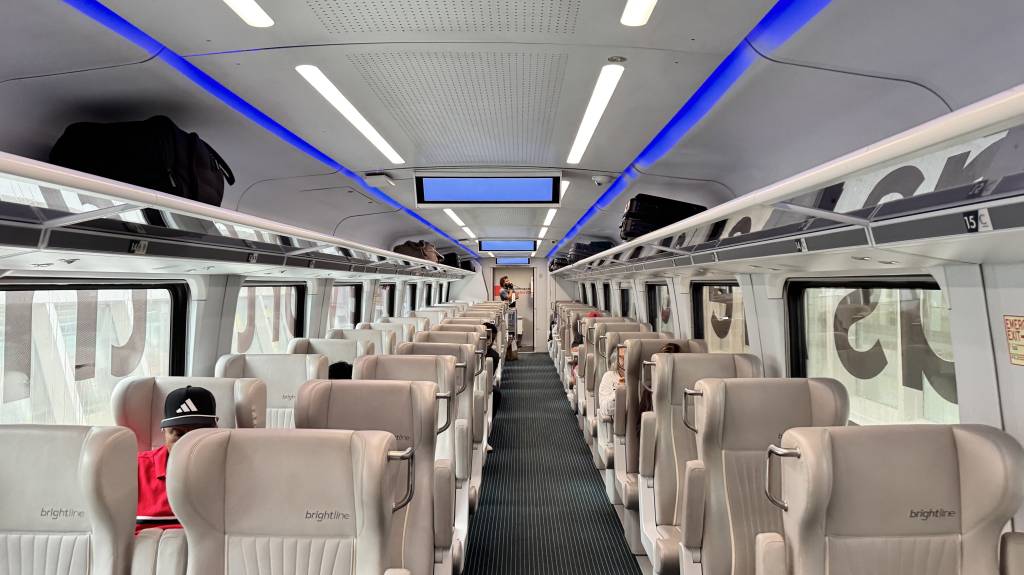 Interior de um trem moderno com assentos bege dispostos em duas fileiras, corredor central e bagageiros superiores. Luzes brancas e azuis iluminam o teto. Passageiros sentados e malas nos bagageiros. Uma pessoa de boné preto e camisa vermelha aparece em primeiro plano. O logo brightline está nos assentos