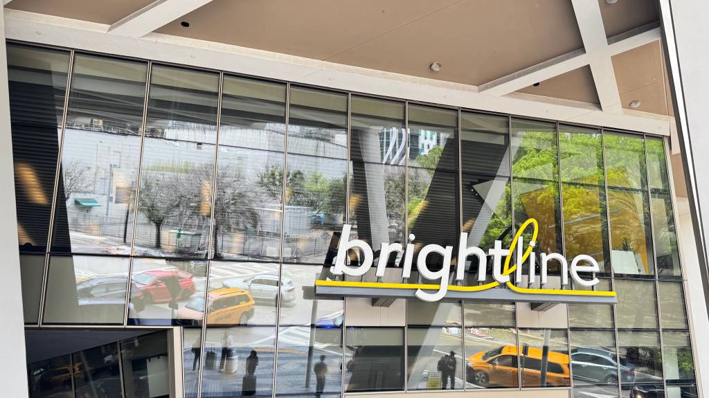 Fachada de vidro de um prédio com o letreiro brightline em branco e amarelo, refletindo carros, árvores e pessoas na rua