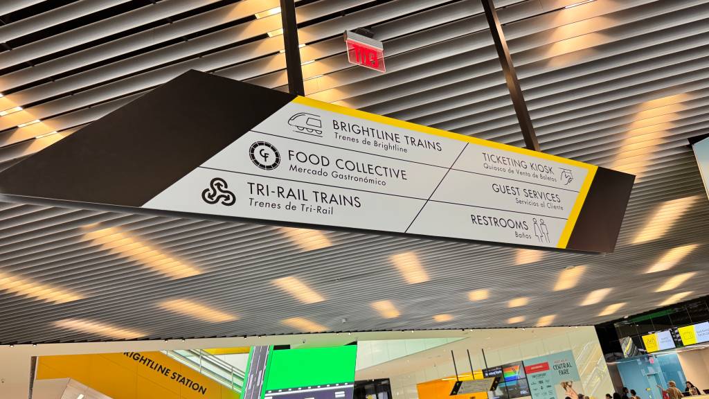 Placa de sinalização suspensa em um terminal, indicando direções para Brightline Trains, Food Collective, Tri-Rail Trains, Ticketing Kiosk, Guest Services e Restrooms, com texto em inglês e espanhol