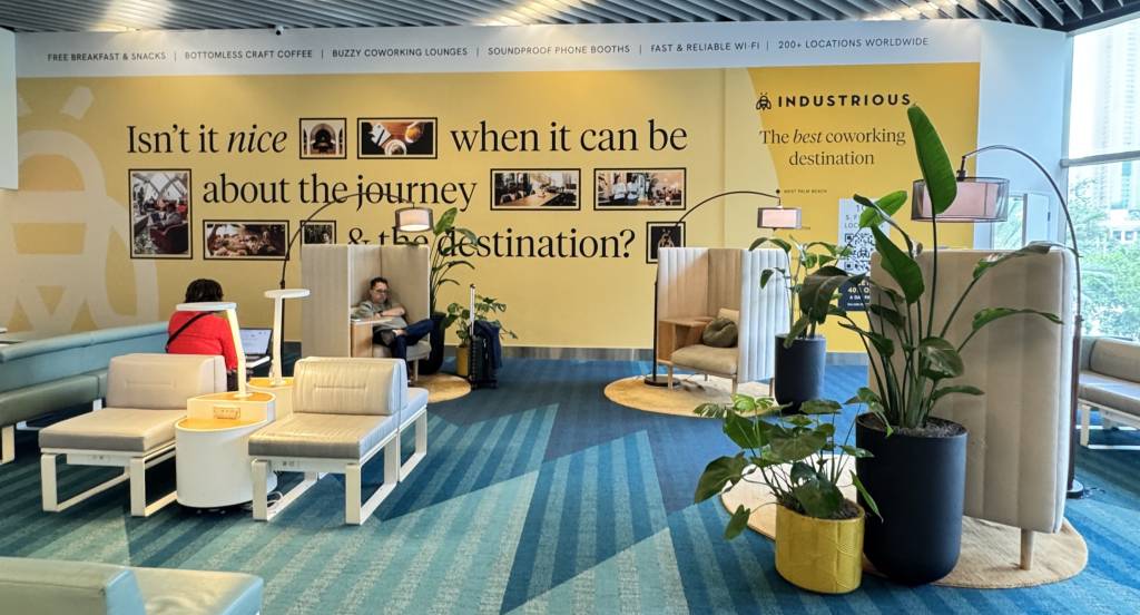 Pessoas relaxando em um lounge de coworking com poltronas e sofás modernos. Uma mulher de blusa vermelha usa um laptop, enquanto um homem de óculos descansa em uma poltrona. Plantas grandes decoram o ambiente, e a parede amarela exibe a frase Isnt it nice when it can be about the journey & the destination? com fotos. O chão tem um tapete azul com padrões geométricos. No alto, um banner lista serviços como FREE BREAKFAST & SNACKS e FAST & RELIABLE WI-FI. À direita, um logo INDUSTRIOUS e The best coworking destination.