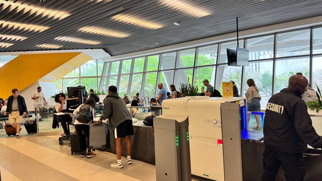 Pessoas em um saguão de aeroporto moderno, com um segurança de costas em primeiro plano, próximo a um scanner de bagagens Orion 920 CX. Ao fundo, passageiros aguardam, alguns sentados, outros em pé, com malas. Grandes janelas revelam árvores e prédios externos, e um teto com iluminação linear completa o ambiente.