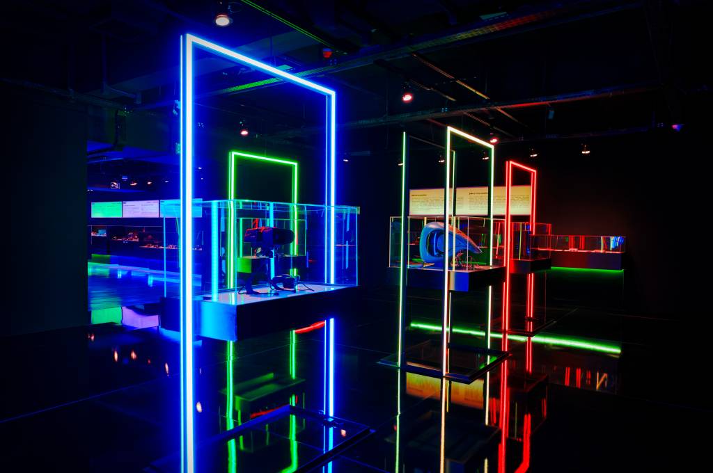 Exposição futurista com salas escuras e displays iluminados por luzes neon azuis, verdes e vermelhas. Em primeiro plano, um display azul com um headset de realidade virtual e um controle. Ao fundo, outros displays com objetos tecnológicos e textos informativos. O chão reflete as luzes, criando um ambiente imersivo