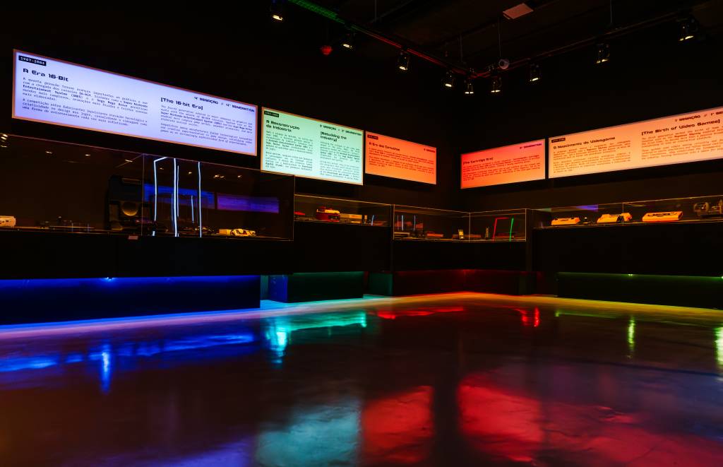 Exposição de videogames com painéis informativos iluminados em branco, verde e laranja, sobre a história dos jogos. Vitrines escuras exibem consoles e acessórios, enquanto o chão reflete luzes coloridas em tons de azul, verde e vermelho, criando um ambiente imersivo