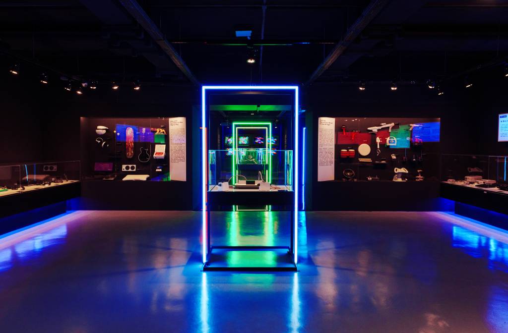 Exposição futurista com luzes neon azuis e verdes, destacando um display central com objetos tecnológicos e painéis informativos nas paredes, refletindo no chão brilhante