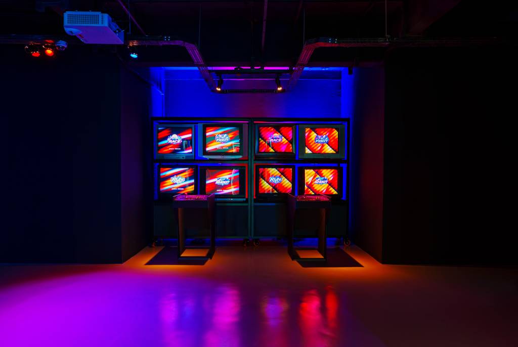 Oito monitores antigos exibem telas de jogos coloridas com as palavras Race, Fight, Run e Shoot, em um ambiente escuro iluminado por luzes neon azuis, roxas e vermelhas. Dois pedestais com controles de arcade estão à frente dos monitores, sugerindo uma área de jogos retrô