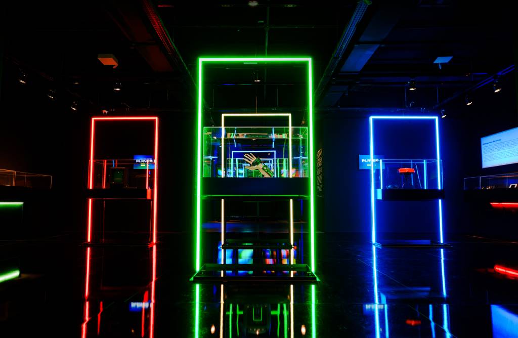 Exposição futurista com três estandes iluminados por neon: vermelho, verde e azul. O estande verde central exibe uma mão robótica dentro de uma caixa de vidro, refletindo no chão escuro.