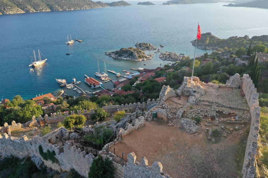 Vista aérea de um porto turco com barcos ancorados em águas azuis, casas com telhados vermelhos e ruínas de um castelo de pedra com uma bandeira da Turquia tremulando no topo