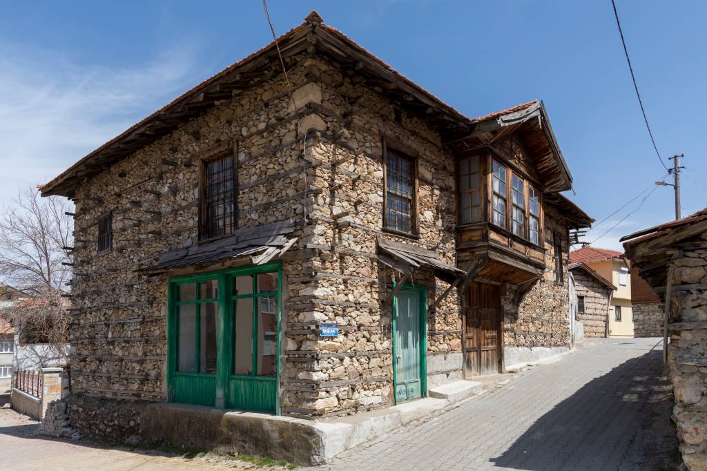 Casa antiga de pedra e madeira, com telhado de telhas e janelas escuras. A fachada principal tem uma porta e uma grande janela verde, e um balcão de madeira no andar superior. Uma rua de paralelepípedos passa ao lado, com outras construções ao fundo sob céu azul