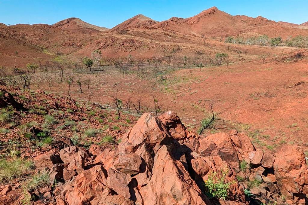 Paisagem árida com montanhas de terra vermelha sob céu azul claro. O primeiro plano mostra rochas avermelhadas e vegetação rasteira esparsa. Ao fundo, colinas e vales com poucas árvores secas e arbustos, destacando a cor vibrante do solo.
