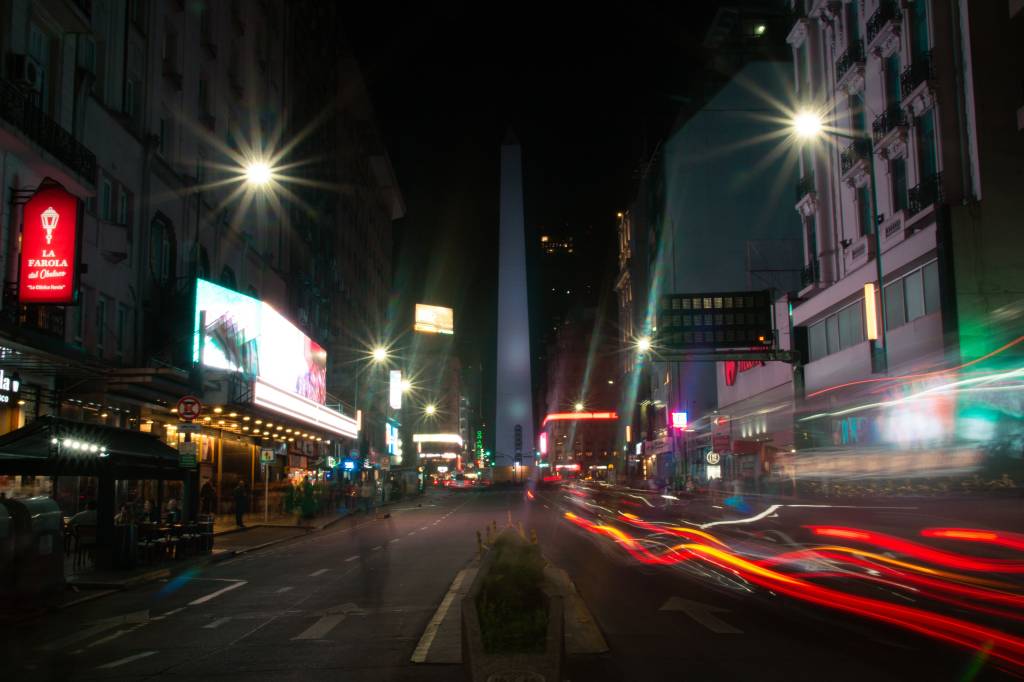 Rua movimentada à noite com o Obelisco de Buenos Aires ao fundo. Prédios iluminados por letreiros e postes de luz, com rastros de luz vermelha e branca de veículos em movimento, indicando longa exposição. Pessoas borradas caminham nas calçadas.