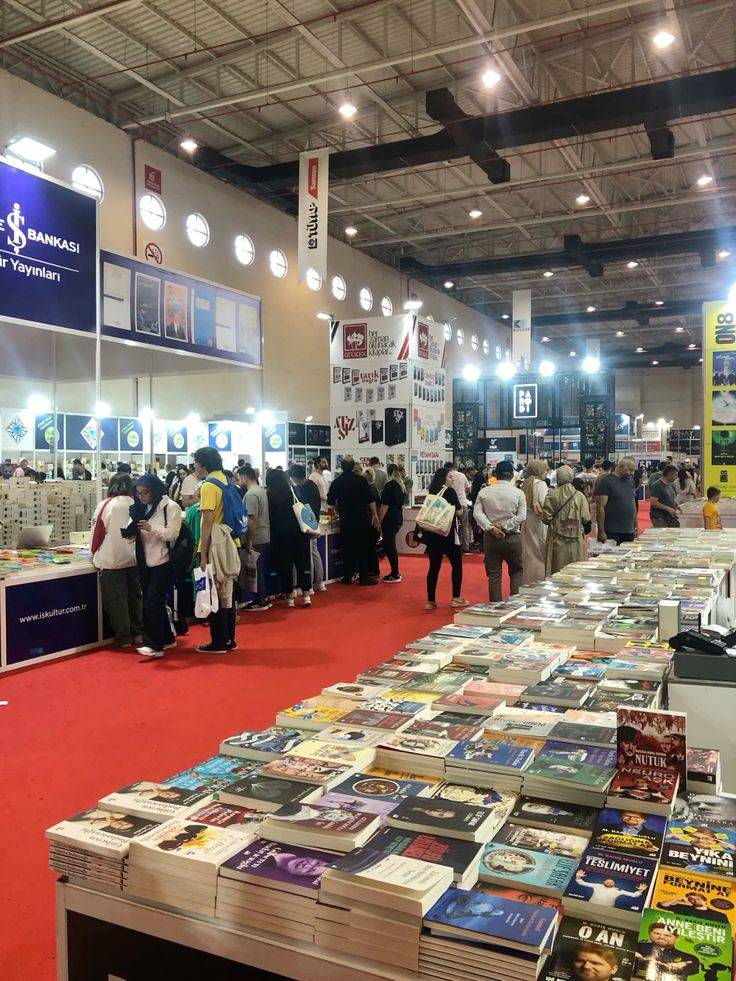 Pessoas circulam em um salão de exposição com carpete vermelho, onde mesas exibem pilhas de livros. Ao fundo, estandes de editoras com banners e cartazes, e luzes no teto.
