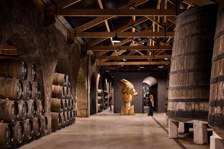 Adega de vinhos com paredes de pedra, barris empilhados à esquerda e ao fundo, e grandes tonéis de madeira à direita. No centro, uma escultura abstrata de madeira e uma mulher de vestido preto observa a obra. O teto exibe vigas de madeira expostas e luminárias pendentes, criando um ambiente rústico e artístico