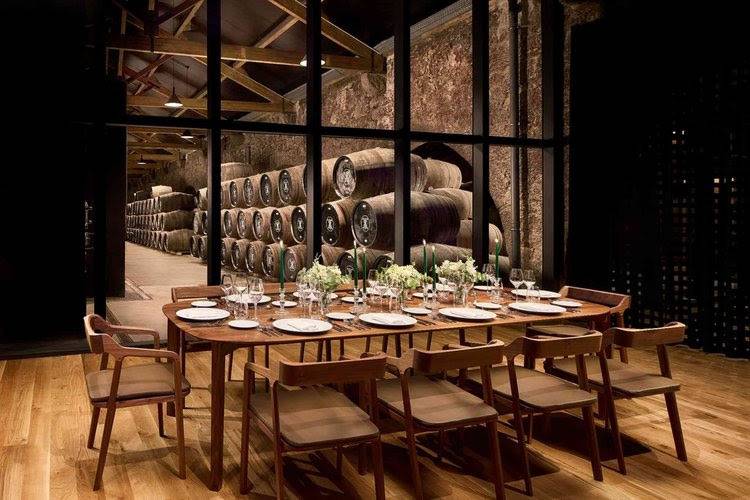 Mesa de jantar retangular de madeira com cadeiras, posta com pratos, copos e arranjos florais, em um ambiente com piso de madeira e parede de vidro que revela uma adega com barris de vinho empilhados