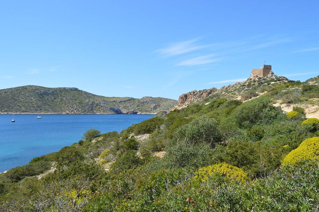 Paisagem costeira com mar azul, duas embarcações e colinas verdes. No topo da colina direita, um castelo de pedra antigo com uma bandeira. Céu azul claro com poucas nuvens.