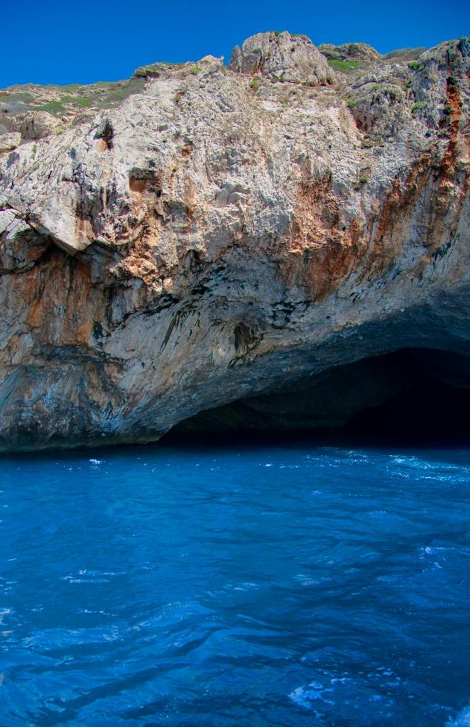 Uma caverna marinha escura com uma abertura em arco na base de um penhasco rochoso, com tons de cinza e laranja, sobre águas azuis intensas e onduladas sob um céu azul claro. Foto sob licença CC BY 2.0
