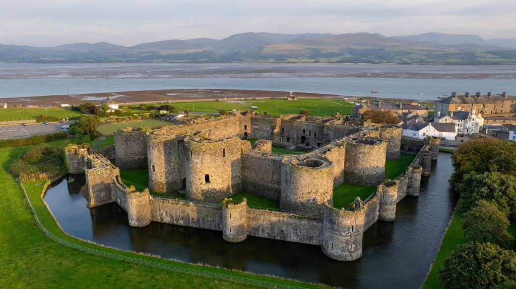 Castelo medieval de pedra com muralhas e torres circulares, cercado por um fosso com água escura e gramado verde. Ao fundo, um estuário com areia exposta e montanhas distantes sob céu claro. Casas e árvores complementam a paisagem