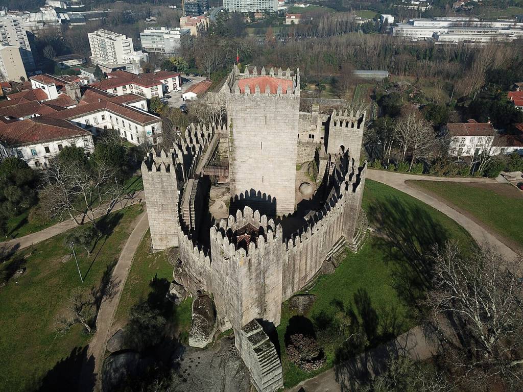 Vista aérea do Castelo de Guimarães, Portugal, com suas muralhas de pedra cinza e ameias pontiagudas. O castelo tem uma torre central mais alta com telhado avermelhado. Ao redor, há gramados verdes, árvores sem folhas e caminhos de terra. Ao fundo, edifícios urbanos e áreas arborizadas completam a paisagem ensolarada. Foto sob licença CC BY-SA 4.0