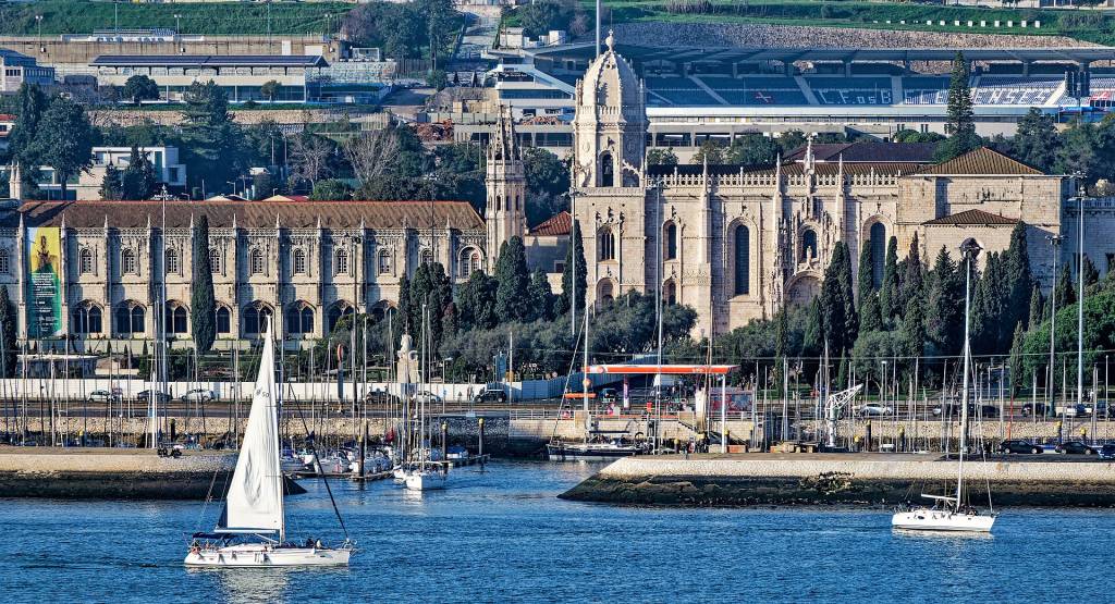Vista panorâmica de Lisboa com o Mosteiro dos Jerónimos em destaque, barcos à vela no rio Tejo e o Estádio do Restelo ao fundo. O mosteiro, em pedra clara, possui arquitetura manuelina com torres e detalhes ornamentados. Um veleiro branco com a vela içada navega em primeiro plano, enquanto outros barcos estão ancorados na marina. A paisagem urbana se mistura com a natureza, sob um céu azul claro