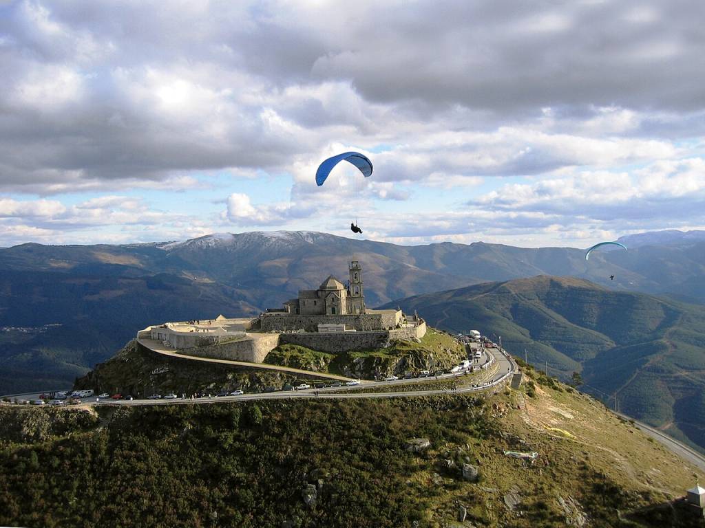 Dois parapentes azuis sobrevoam um santuário de pedra no topo de uma montanha, com uma estrada sinuosa e carros estacionados. Ao fundo, montanhas verdes e picos nevados sob um céu nublado com aberturas azuis