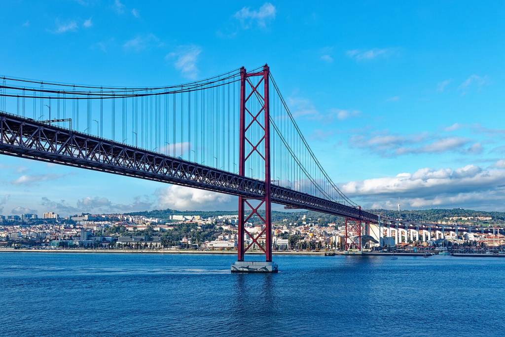 Ponte 25 de Abril em Lisboa, Portugal, com sua estrutura vermelha e cinza sobre o rio Tejo azul, sob um céu claro com nuvens esparsas. A margem da cidade, com edifícios e vegetação, aparece ao fundo