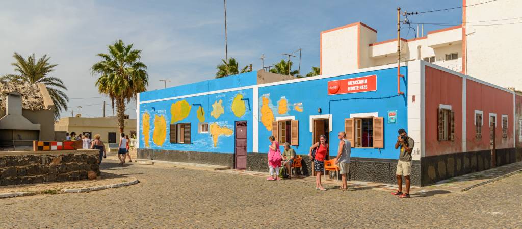 Rua de paralelepípedos com pessoas caminhando e conversando em frente a uma mercearia azul com mapas amarelos pintados na parede e um prédio vermelho ao lado, sob céu azul claro