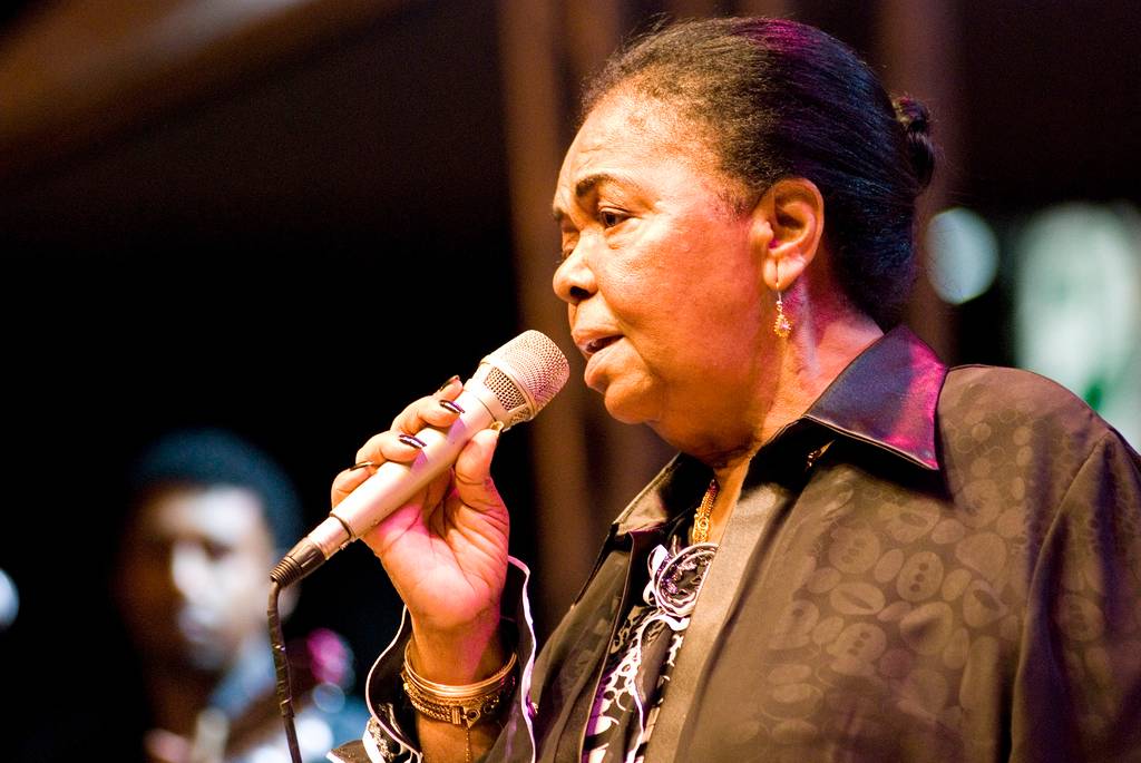 Cantora negra de meia-idade com cabelo preso, usando brincos dourados e blusa preta estampada, segurando um microfone prateado com a mão direita e cantando em um palco escuro