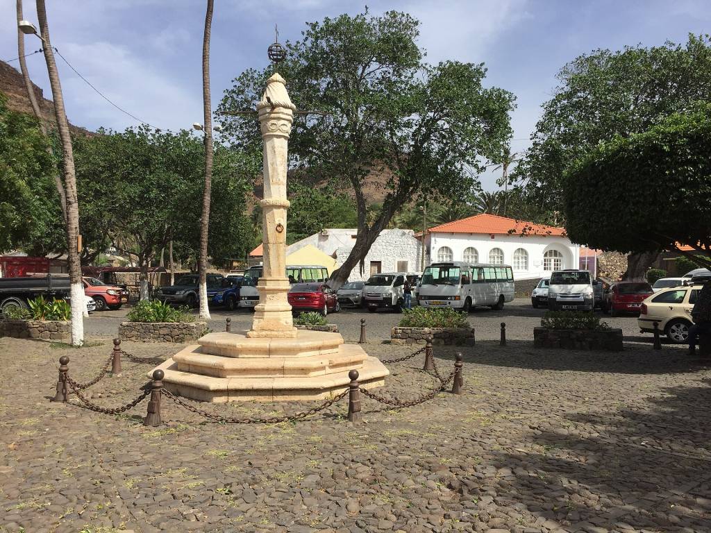 Praça de paralelepípedos com um pelourinho de pedra no centro, cercado por correntes. Ao fundo, carros e vans estacionados, casas brancas com telhados de terracota e árvores frondosas sob céu parcialmente nublado
