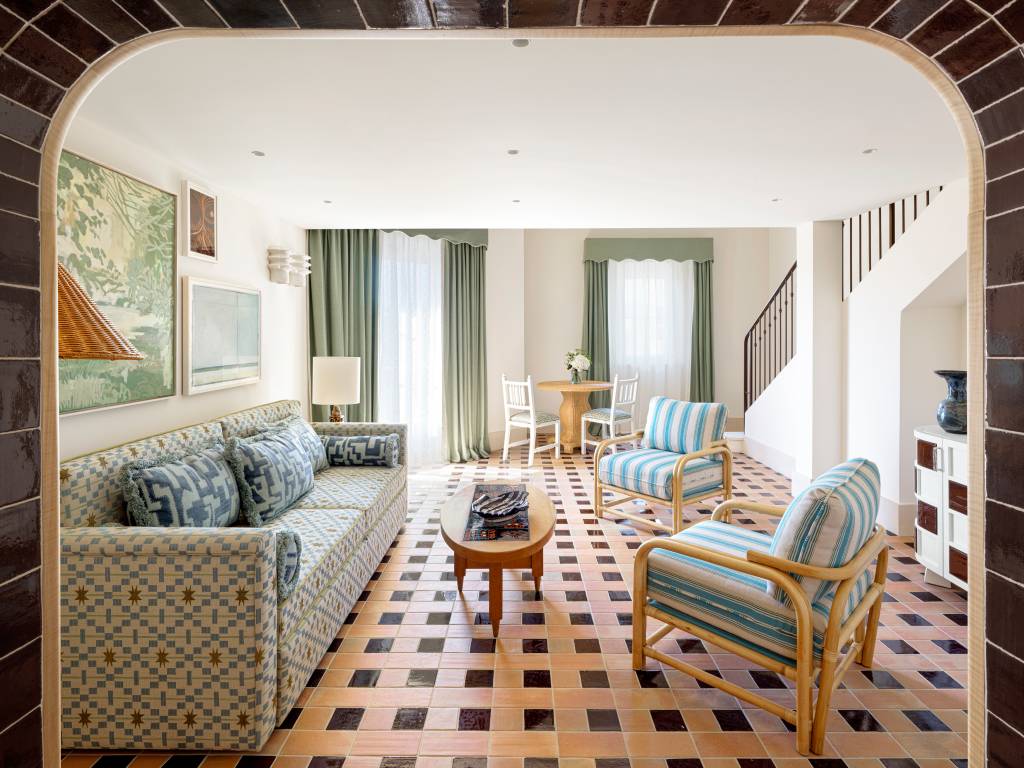 Sala de estar com sofá estampado em azul e bege, duas poltronas de bambu com estofado listrado azul e branco, mesa de centro oval de madeira escura, e piso de ladrilhos marrons e beges. Ao fundo, uma mesa redonda com cadeiras brancas e uma escada com corrimão preto.