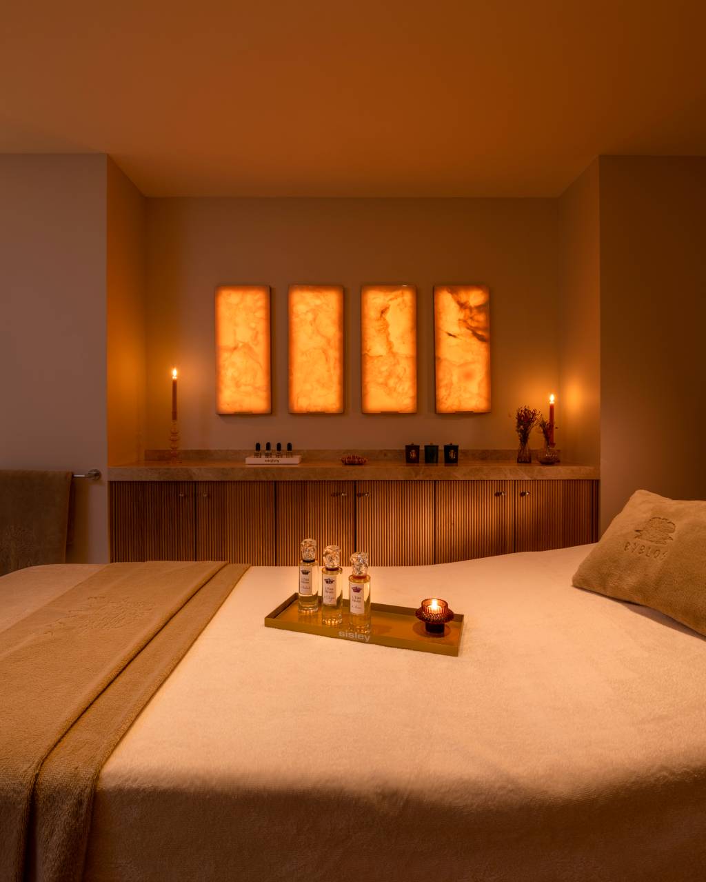 Cama de massagem coberta por lençol branco e manta bege, com bandeja dourada contendo três frascos de perfume e uma vela acesa. Ao fundo, uma bancada de madeira com quatro painéis iluminados de ônix laranja e duas velas em castiçais, criando um ambiente relaxante e acolhedor