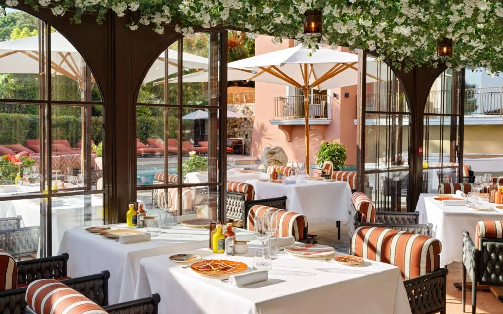 Restaurante luxuoso com mesas brancas e cadeiras listradas em ambiente envidraçado, decorado com flores brancas no teto. Ao fundo, uma piscina com espreguiçadeiras e um prédio cor-de-rosa com varandas.