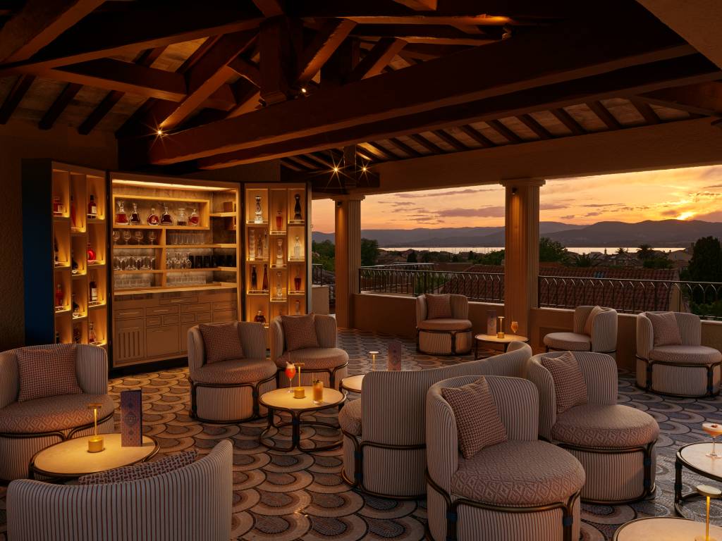 Terraço de bar luxuoso ao entardecer, com teto de madeira e vigas expostas. Há um bar iluminado com garrafas e copos, e várias poltronas estofadas em tons claros, com mesas de centro servindo coquetéis. Ao fundo, um pôr do sol vibrante sobre um lago e montanhas