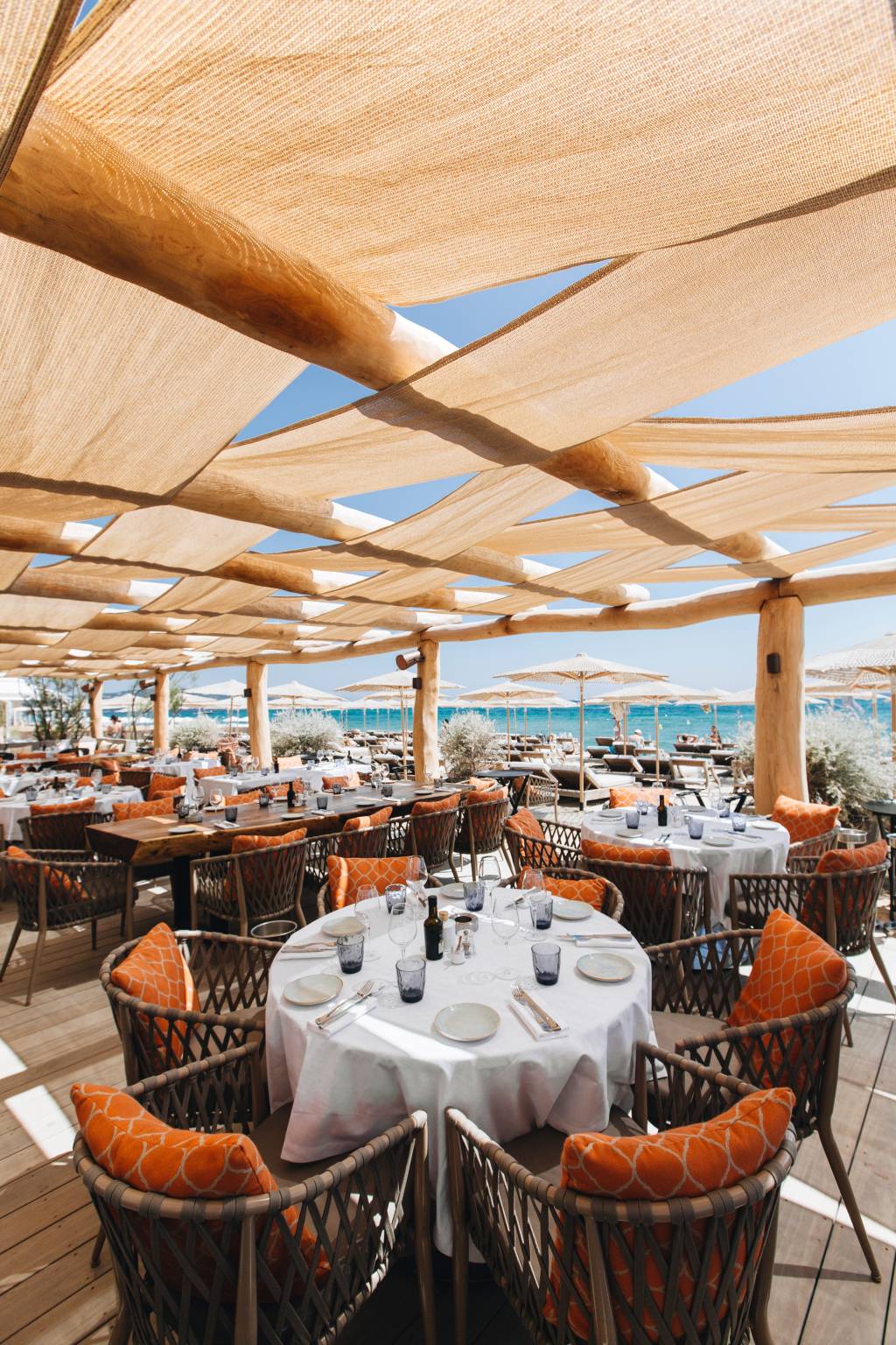 Restaurante à beira-mar com mesas redondas e retangulares, cadeiras de vime com almofadas laranja, sob um teto de tecido bege. Ao fundo, espreguiçadeiras e guarda-sóis na praia, com o mar azul-turquesa e céu claro