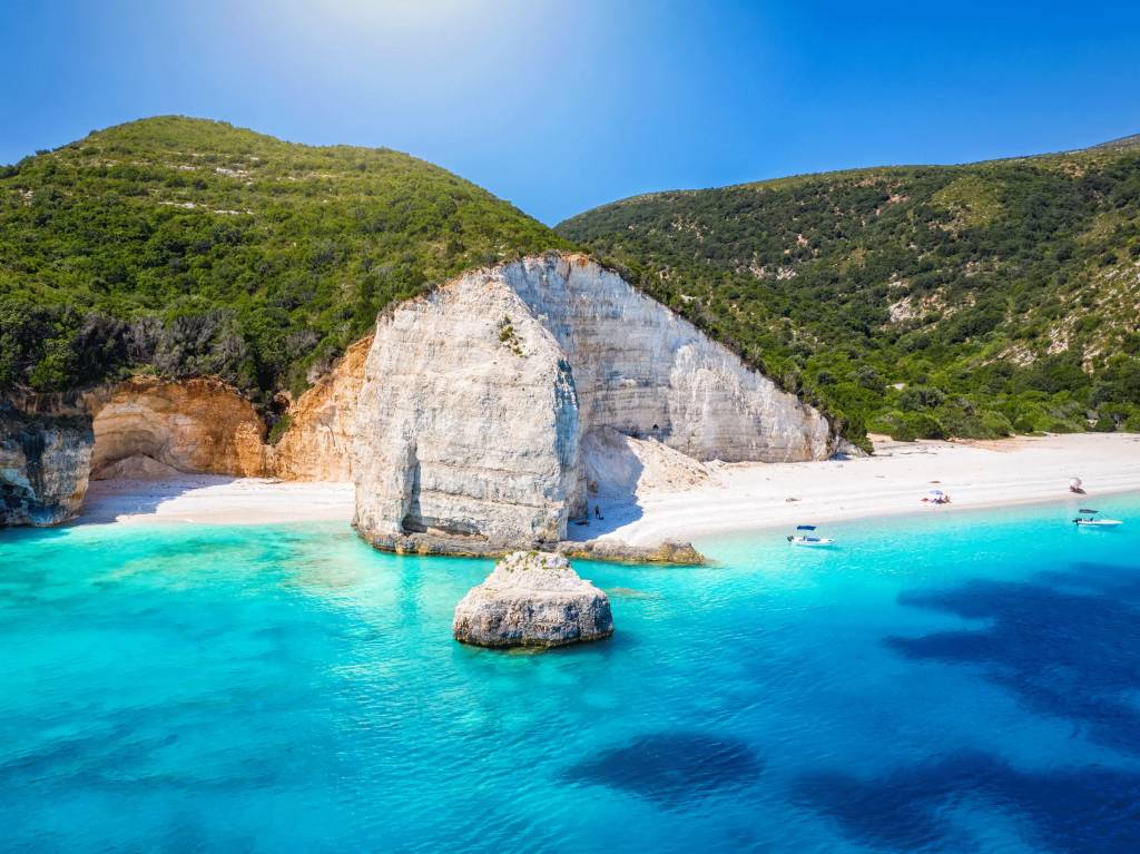 Praia paradisíaca com areia branca, mar azul-turquesa e falésias rochosas. Montanhas verdes cobrem o topo das falésias. Pequenos barcos brancos flutuam na água cristalina, e um rochedo solitário emerge do mar. O céu azul claro completa a paisagem ensolarada