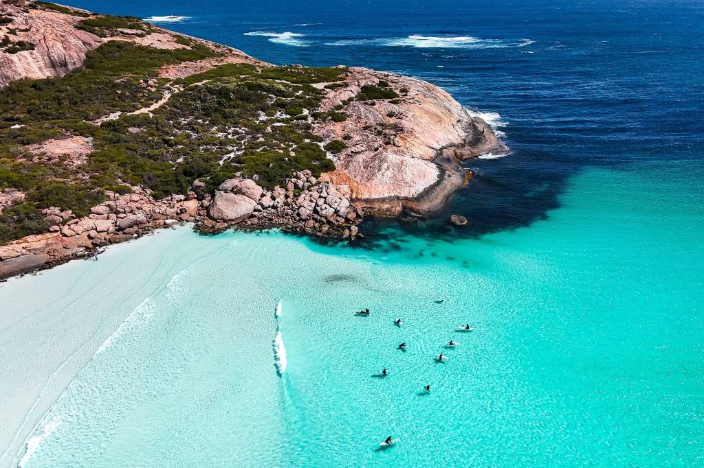Vista aérea de uma praia com areia branca e mar azul-turquesa, onde surfistas esperam ondas. Uma formação rochosa coberta por vegetação verde margeia a praia, e o oceano profundo se estende ao fundo