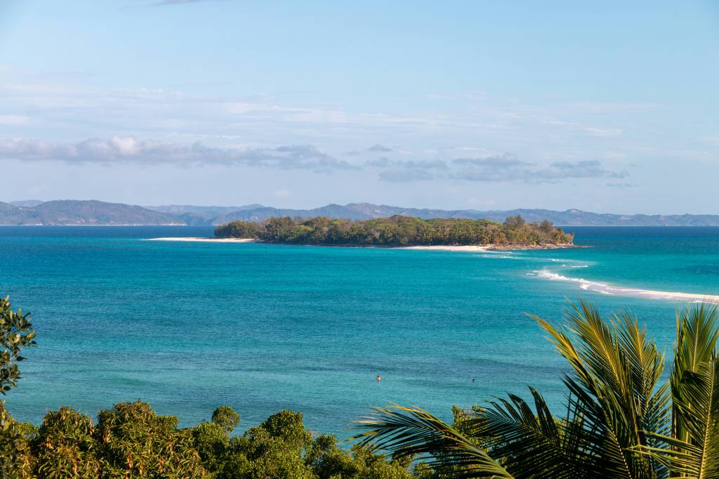 Vista aérea de uma ilha tropical com águas azul-turquesa, faixa de areia branca e vegetação densa. Montanhas distantes no horizonte sob céu azul com nuvens esparsas. Folhas de palmeira em primeiro plano à direita e folhagem verde à esquerda. Duas pessoas nadam na água cristalina