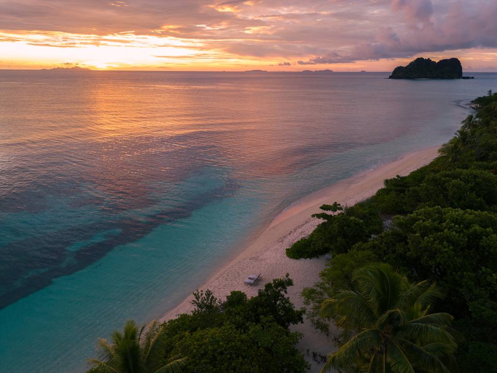 Vista aérea de uma praia tropical ao pôr do sol, com areia clara e vegetação densa à direita. O mar exibe tons de azul turquesa próximo à costa e laranja rosado no horizonte, refletindo o céu com nuvens alaranjadas e roxas. Uma pequena ilha montanhosa escura se destaca à direita no mar.