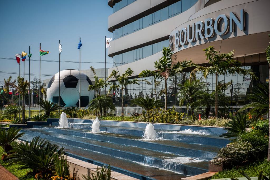 Fachada de um edifício moderno com o nome BOURBON em destaque, com janelas espelhadas refletindo o céu azul. Em frente, uma fonte em cascata com jatos dágua, palmeiras e arbustos. Ao fundo, uma enorme bola de futebol preta e branca e mastros com bandeiras de diversos países