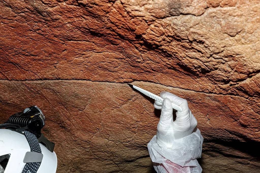 Mão enluvada de branco aponta uma ferramenta branca para uma fina linha escura em uma parede rochosa de tons terrosos, com um capacete branco no canto inferior esquerdo