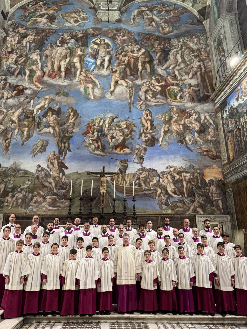 Um coral de homens e meninos em vestes brancas e roxas posa em frente ao afresco O Juízo Final de Michelangelo, na Capela Sistina. No centro, um crucifixo e castiçais.