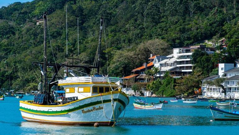 Barco de pesca branco e amarelo com o nome Tundão e Pumba ancorado em águas azuis, com casas e vegetação densa em uma encosta ao fundo