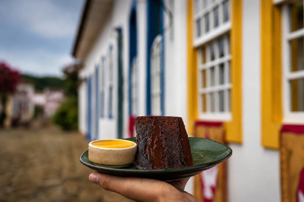 Mão segura um prato verde com um pedaço de bolo de chocolate escuro e uma tigela pequena de molho amarelo, em frente a casas coloniais brancas com janelas azuis e amarelas