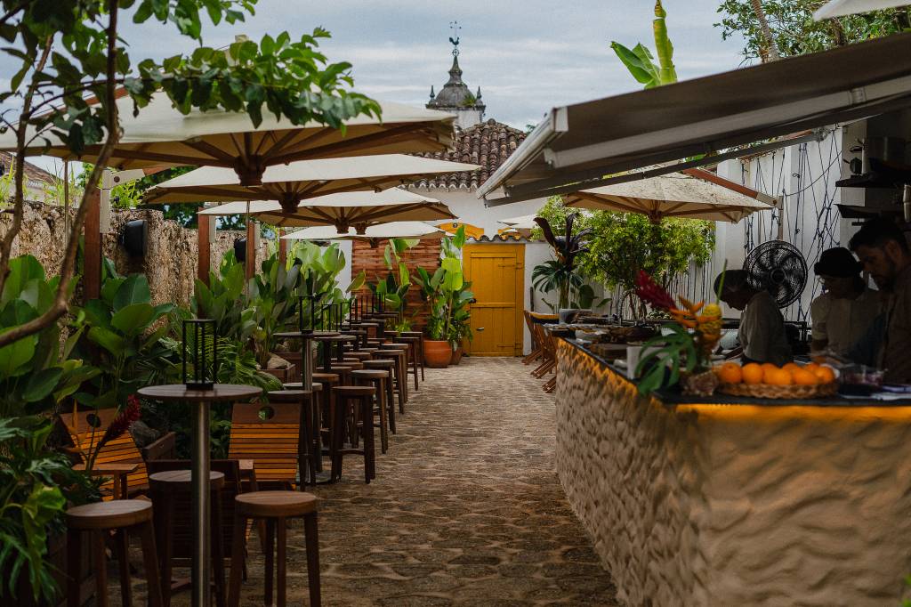 Pátio externo de restaurante com mesas e banquetas de madeira sob guarda-sóis brancos, plantas verdes e um balcão de pedra com frutas e funcionários. Ao fundo, uma porta amarela e um telhado colonial com cata-vento