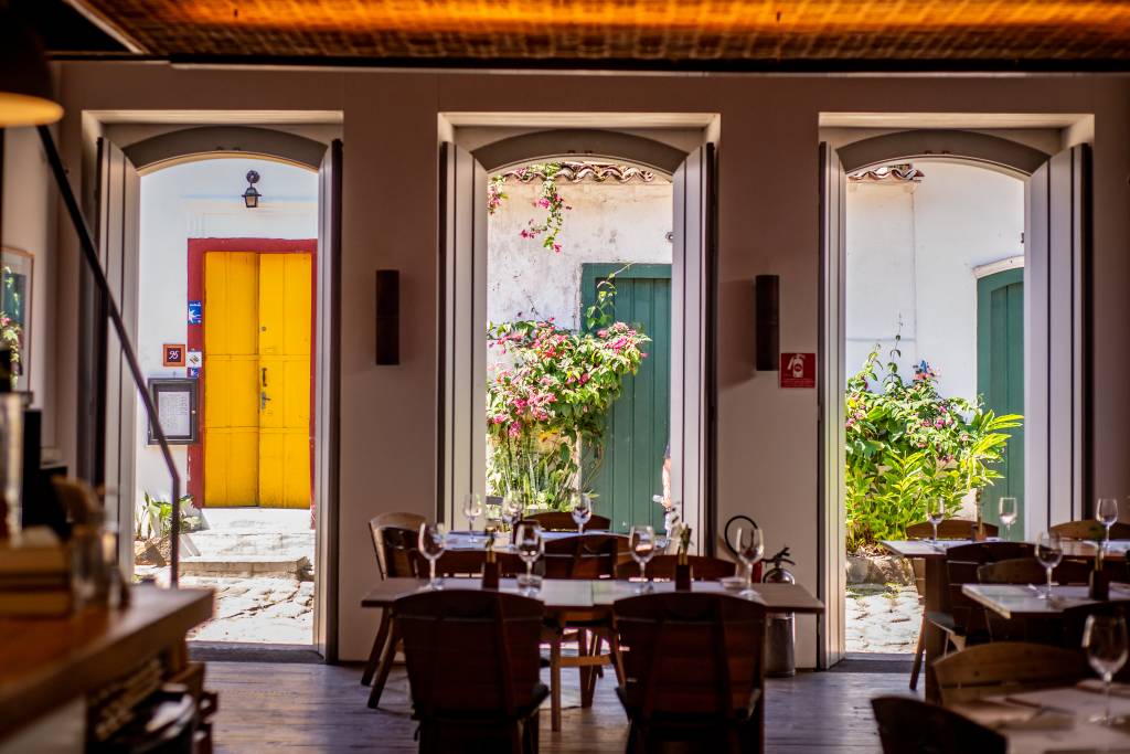 Interior de um restaurante com mesas de madeira e cadeiras escuras, com taças de vinho. Três portas arqueadas revelam um pátio externo com plantas e flores, e uma porta amarela vibrante ao fundo. O chão é de madeira escura e o teto tem um acabamento rústico