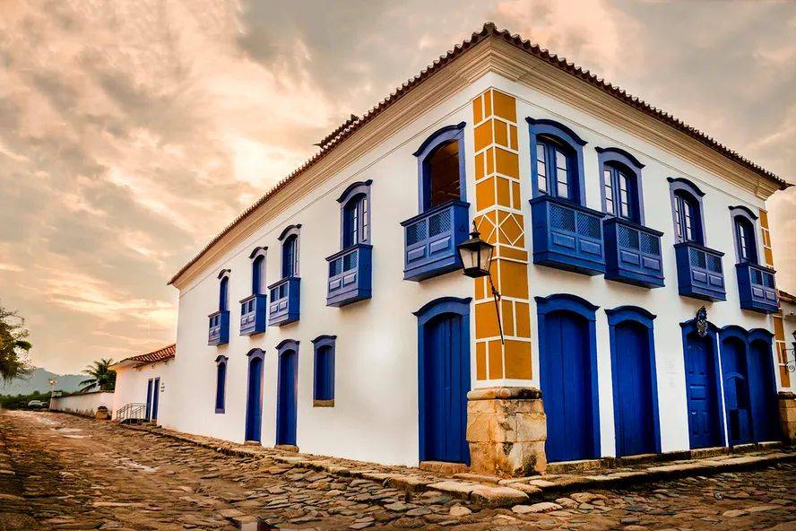 Casarão colonial branco com detalhes em azul e amarelo, telhado de barro, janelas e portas azuis, e sacadas com grades azuis. Um poste de luz antigo está na esquina. A rua de paralelepípedos se estende à esquerda, sob um céu alaranjado com nuvens esparsas