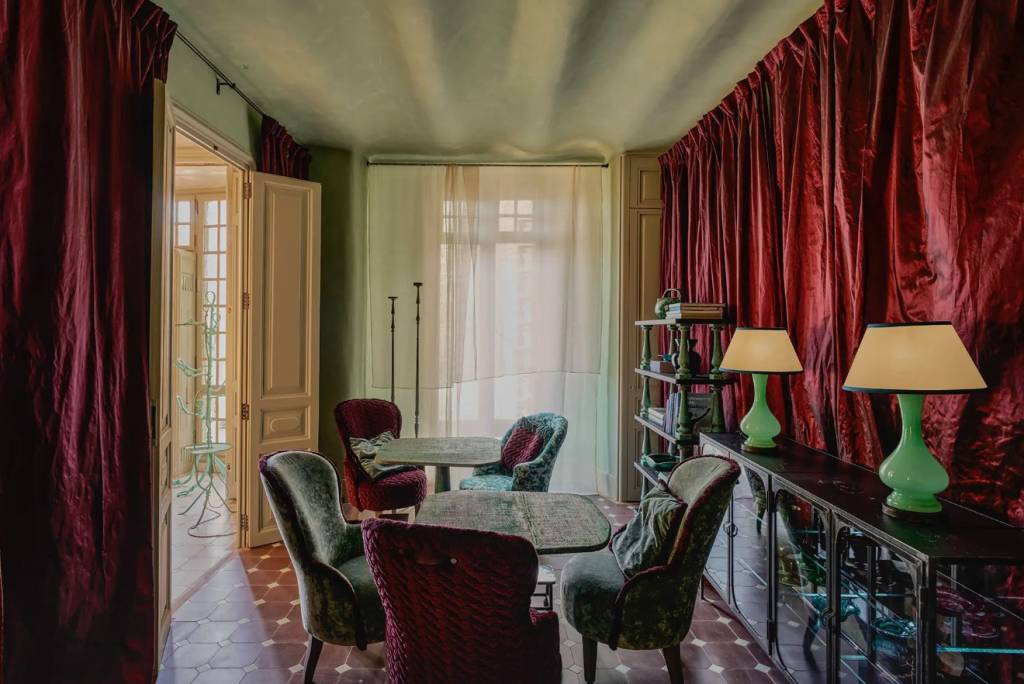 Sala com paredes verde-claras, cortinas de veludo vermelho-escuro e piso de ladrilhos marrons. Há quatro poltronas estofadas em veludo verde e vinho ao redor de duas mesas redondas. À direita, um aparador preto com duas luminárias de abajur creme e base verde-clara. Ao fundo, uma porta aberta revela um ambiente iluminado com janelas e uma estrutura metálica decorativa
