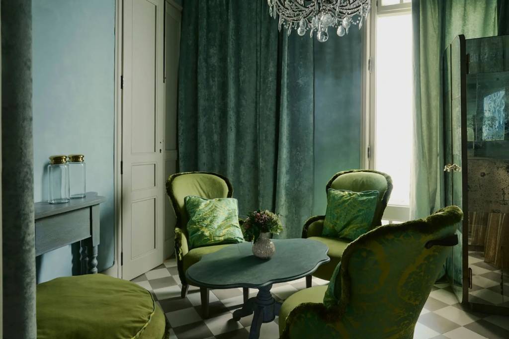 Sala de estar com paredes azul-claras e piso xadrez cinza e branco. Duas poltronas verdes com almofadas estampadas, uma mesa de centro oval com vaso de flores, e um pufe verde. Cortinas verde-escuras em frente a uma janela iluminada, e um lustre de cristal no teto. À esquerda, uma mesa de cabeceira com dois potes de vidro