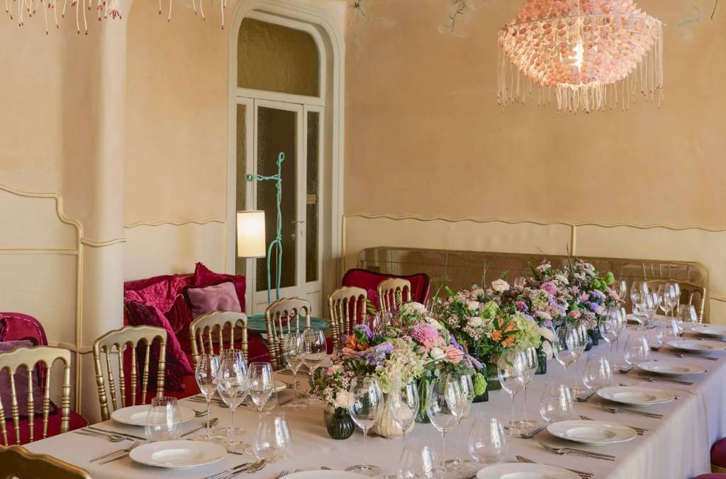 Mesa de jantar longa com toalha branca, pratos, talheres e taças de vinho, decorada com arranjos florais coloridos. Cadeiras douradas e sofás vermelhos ao redor, sob um lustre rosa e uma luminária de chão azul, em um ambiente com paredes bege e janelas altas