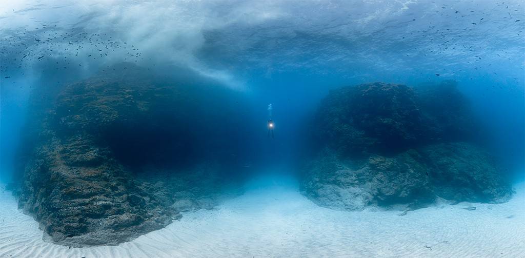 Mergulhador solitário com lanterna ilumina o fundo do mar azul profundo, entre duas grandes formações rochosas escuras e um leito de areia clara. Pequenos peixes nadam perto da superfície.