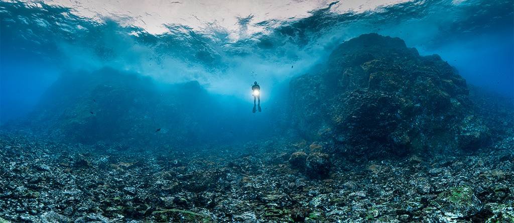 Mergulhador solitário com uma lanterna subaquática brilhante, nadando em águas azuis profundas sobre um leito marinho rochoso e irregular. A superfície da água, acima, reflete a luz do sol em ondas. À direita, uma grande formação rochosa escura se eleva do fundo.